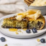 A slice of Blueberry Cornbread on a plate topped with honey butter and honey drizzling over the top.