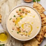 Ultra smooth and creamy hummus in a bowl drizzled with olive oil, dusted with paprika and topped with chickpeas for garnish surrounded by warmed pita triangles.