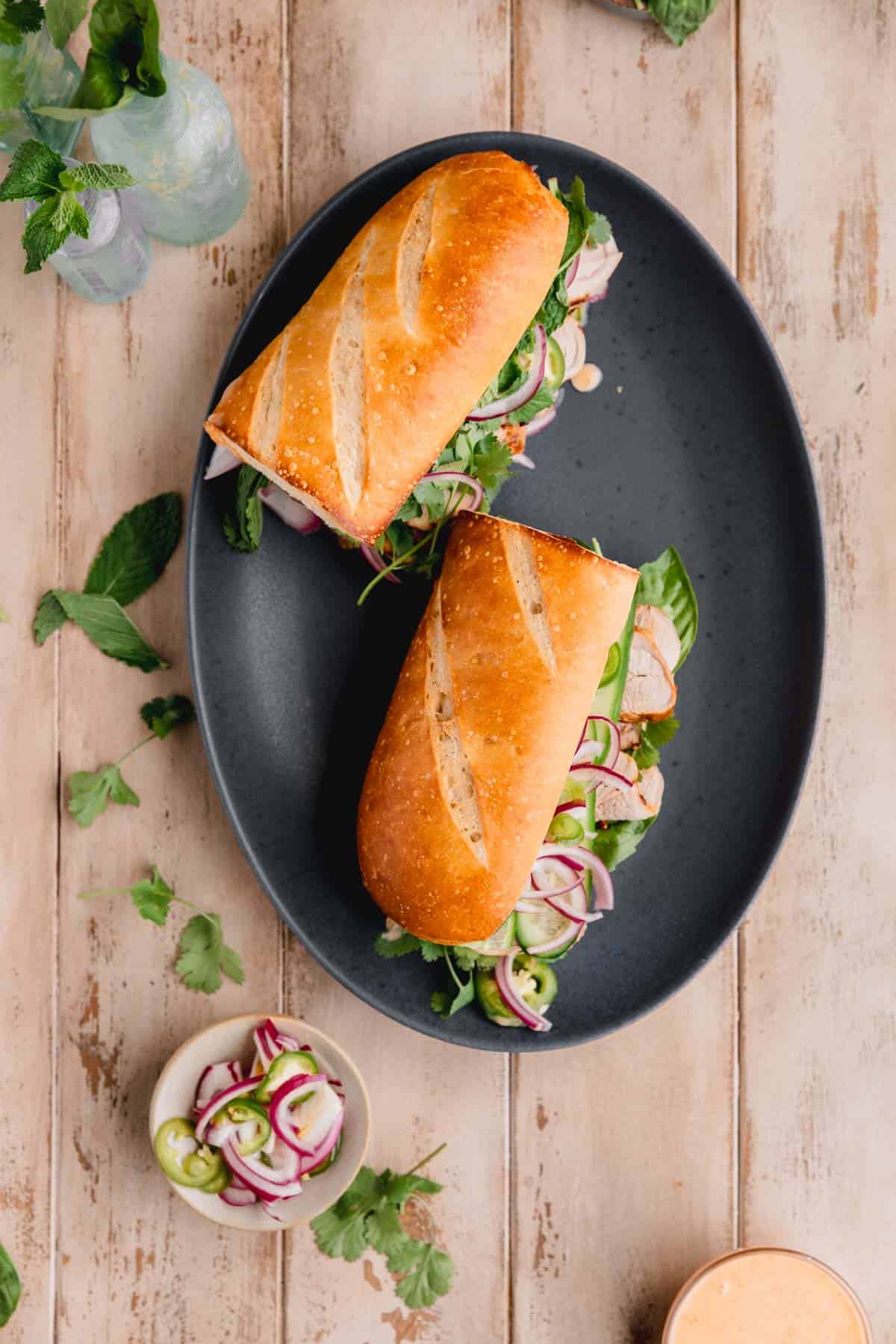 Pork tenderloin baguette on a plate with herbs and pickled veggies to the side. 