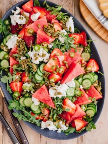 Spicy Watermelon Arugula Salad with a sweet and smoky Chipotle Strawberry Vinaigrette on a large salad platter with serving utensils aside.