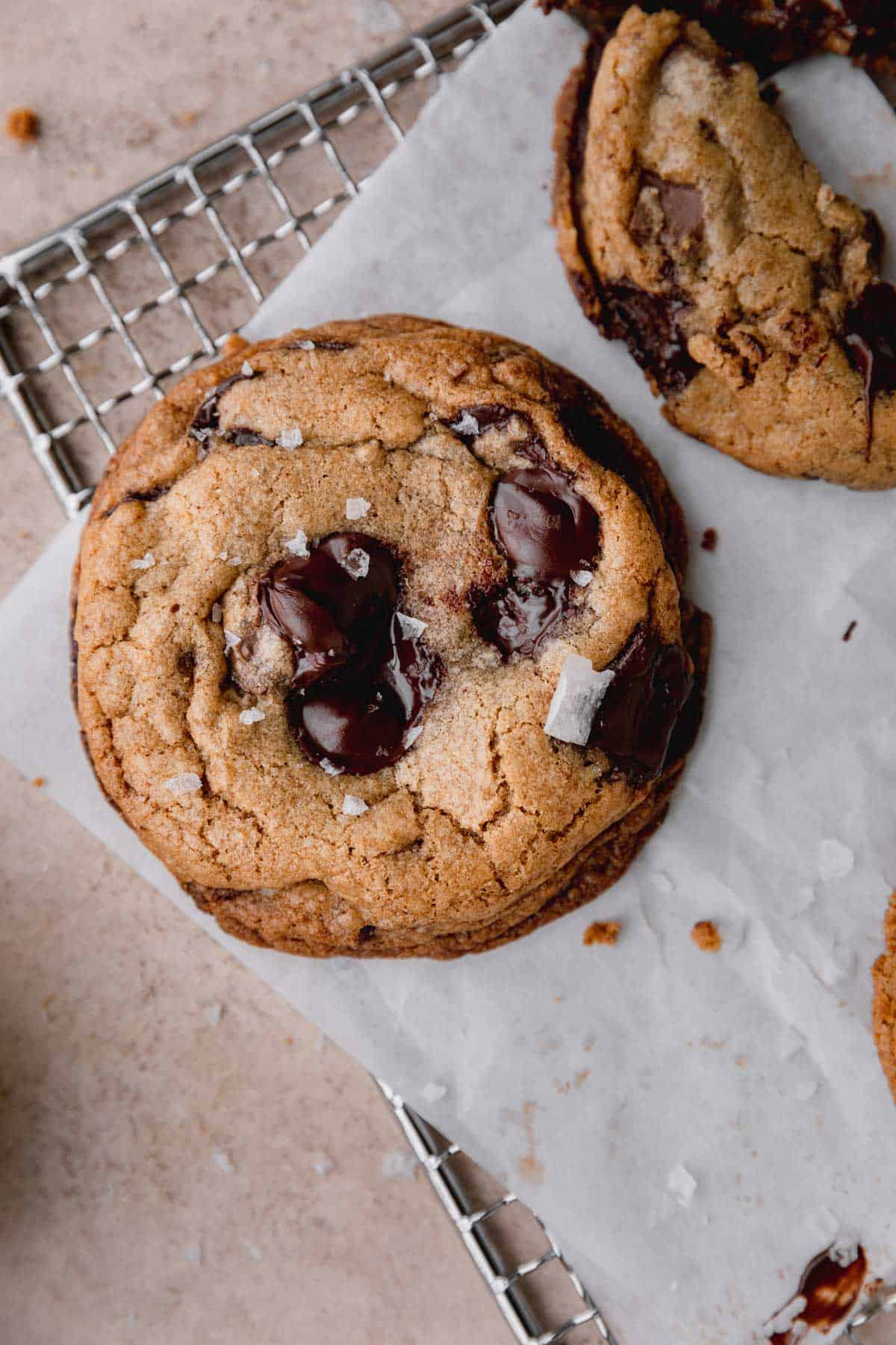 Brown Butter Toffee Chocolate Chip Cookie on a cooling rack with parchment paper, topped with flaky sea salt and melty chocolate.