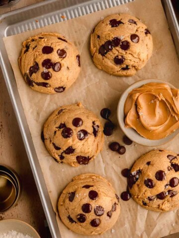 Chocolate Chip Peanut Butter Cookies on a sheet tray lined with parchment paper, topped with flaky sea salt and a dish of peanut butter next to them.