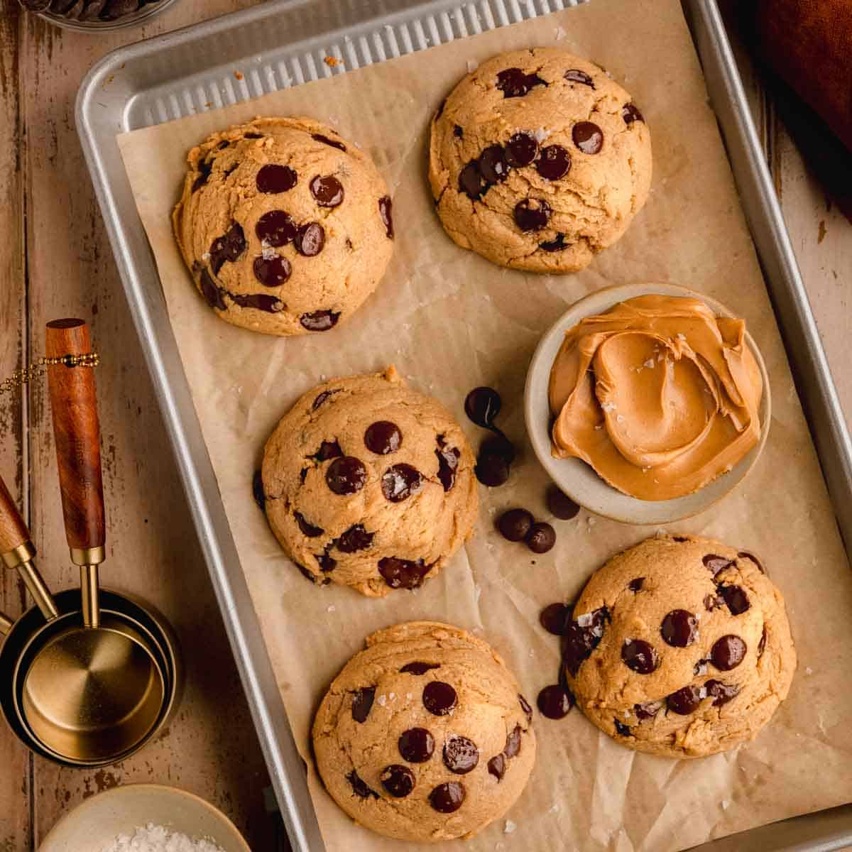 Chocolate Chip Peanut Butter Cookies on a sheet tray lined with parchment paper, topped with flaky sea salt and a dish of peanut butter next to them.