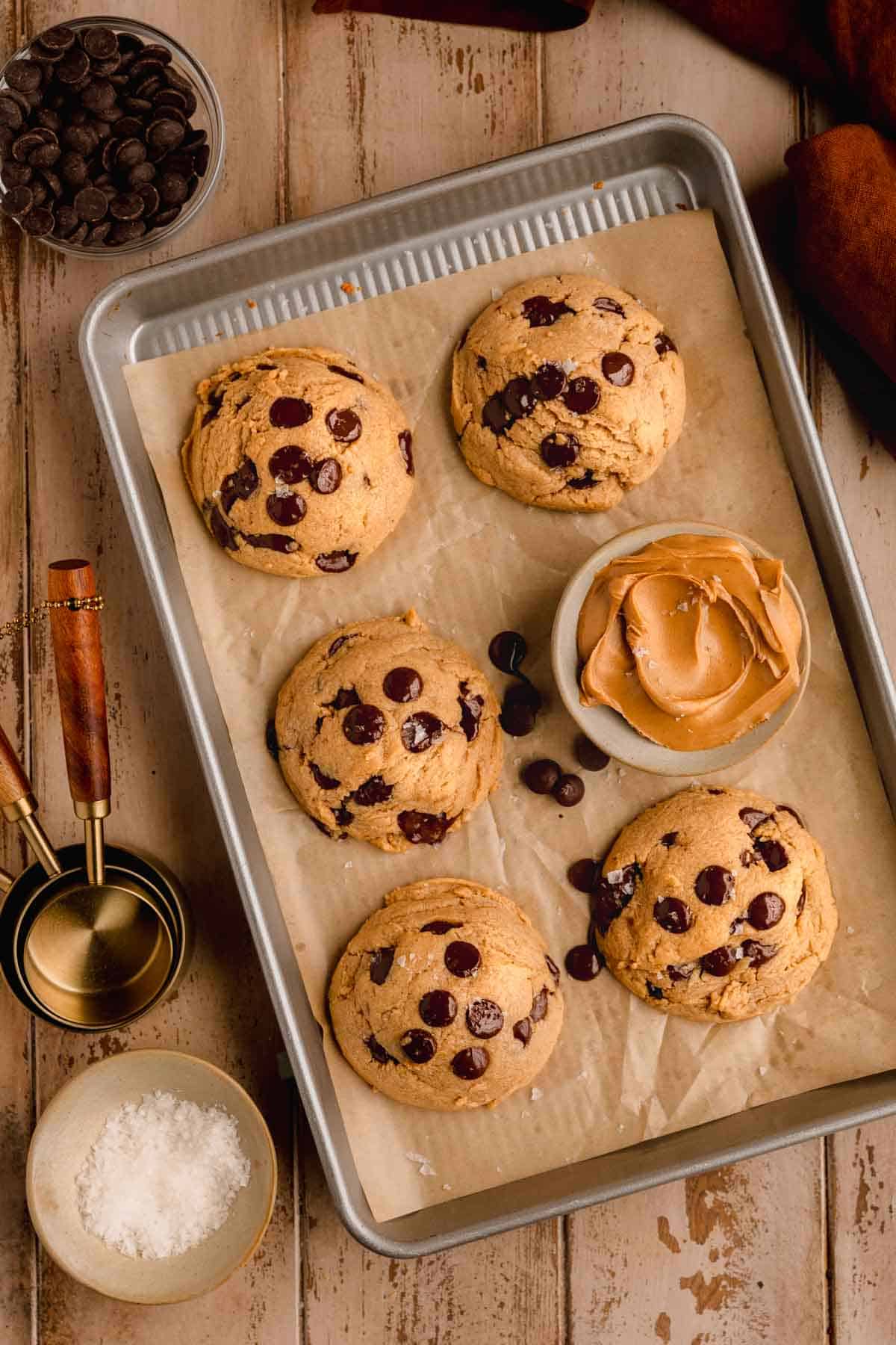 Chocolate Chip Peanut Butter Cookies on a sheet tray lined with parchment paper with a dish of peanut butter and dish of salt as well as measuring cups and chocolate chips to the side. 