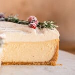 Ultra creamy Eggnog Cheesecake topped with eggnog whipped cream and decorated with sugared cranberries and rosemary with a slice cut out so you can see the layers.