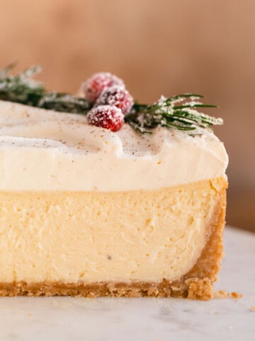 Ultra creamy Eggnog Cheesecake topped with eggnog whipped cream and decorated with sugared cranberries and rosemary with a slice cut out so you can see the layers.