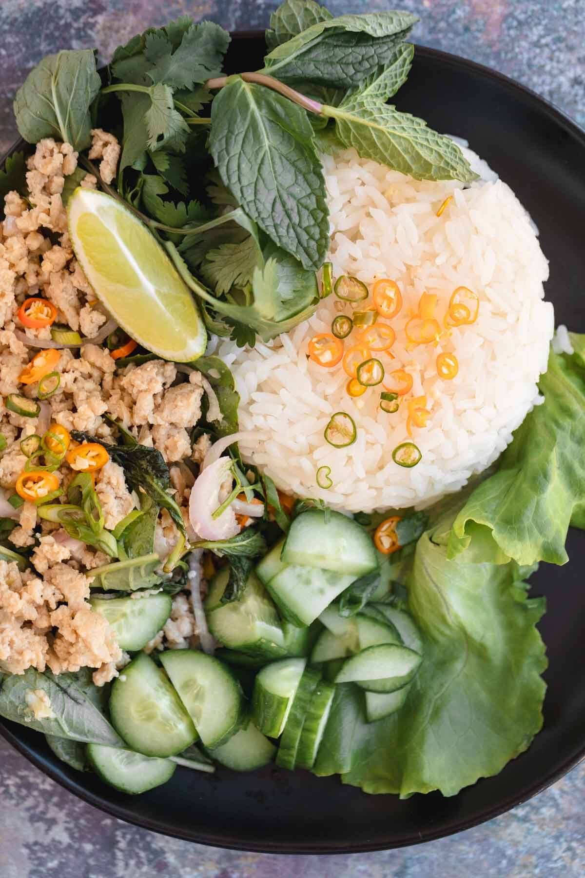 Chicken Larb with Jasmine rice, Thai chile peppers, cucumber and herbs on a plate with a lime wedge.