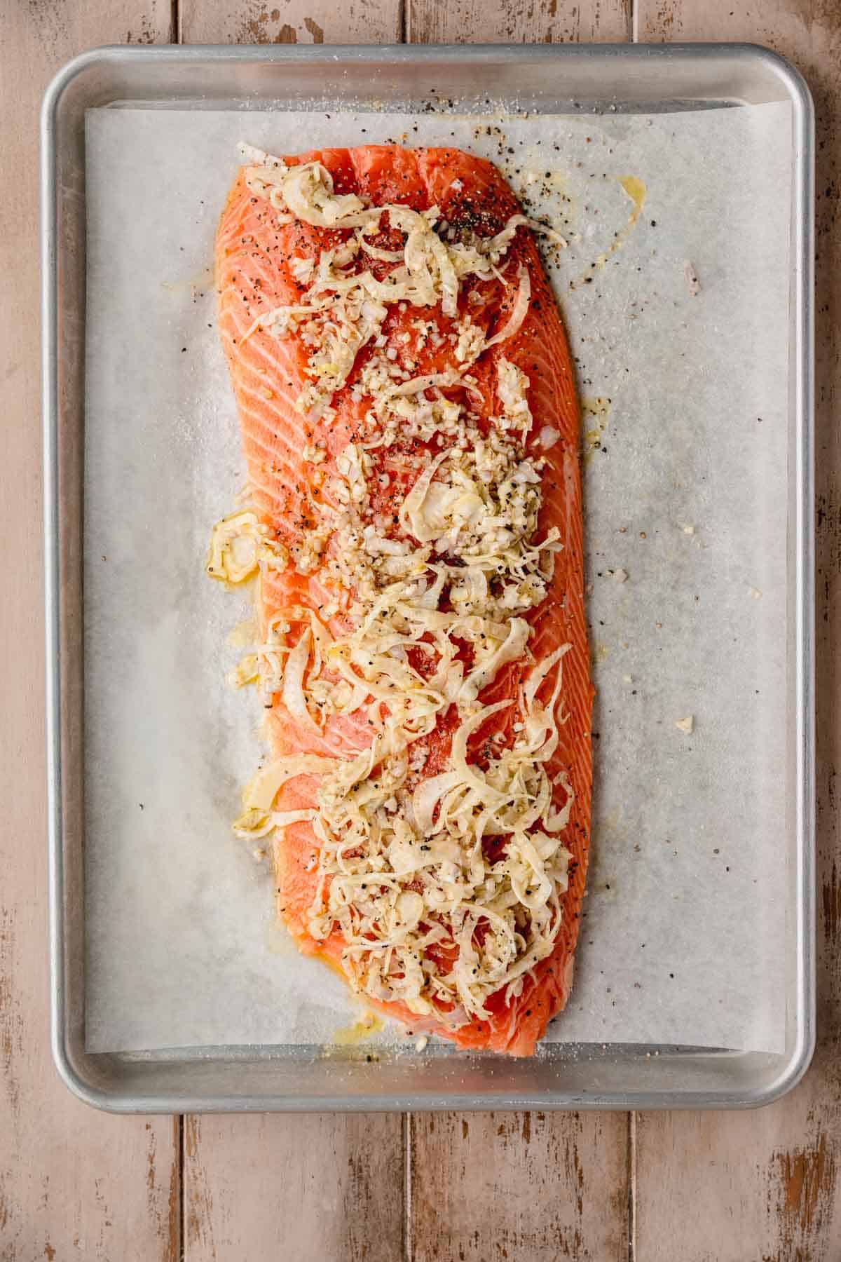 A salmon filet on a sheet tray topped with a fennel and garlic mixture.