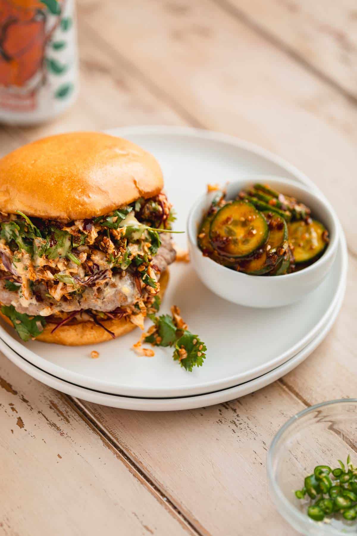 Pork Dumpling Burger on a plate with a dish of spicy cucumbers and a drink on the table.